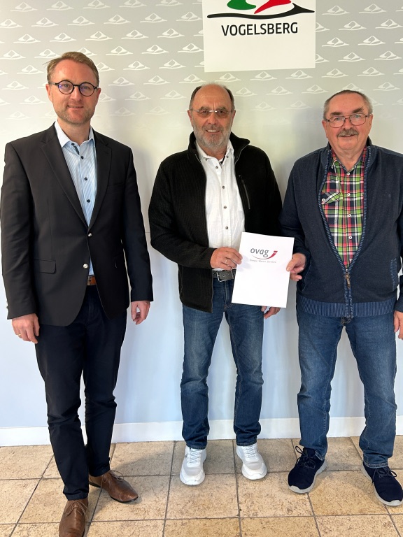 Drei Männer stehen vor einer Fotowand und präsentieren einen Spendenbescheid