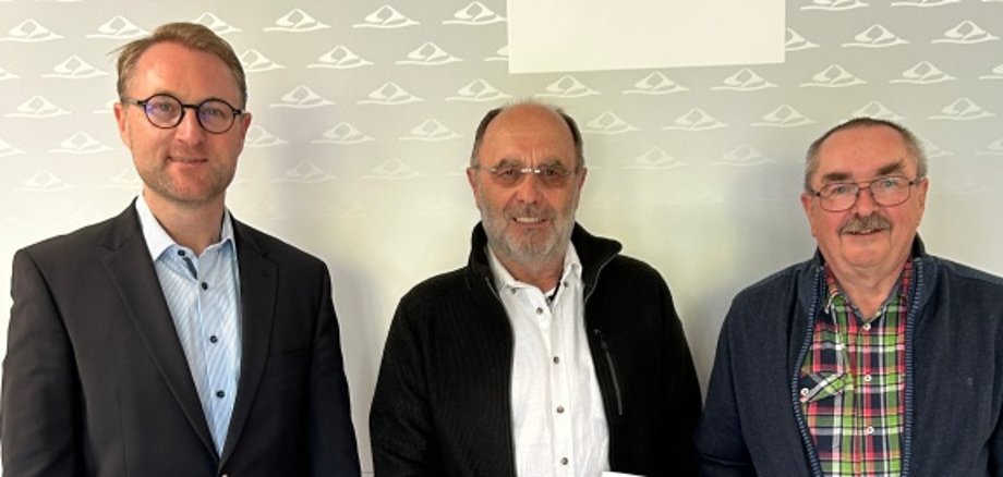 Drei Männer stehen vor einer Fotowand und präsentieren einen Spendenbescheid