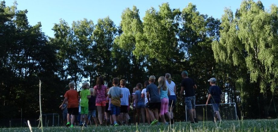 Kinder laufen in einer Reihe über einen Sportplatz. Im Huntergrund sind Bäume zu sehen, die Kinder zeigen den Rücken zur Kamera