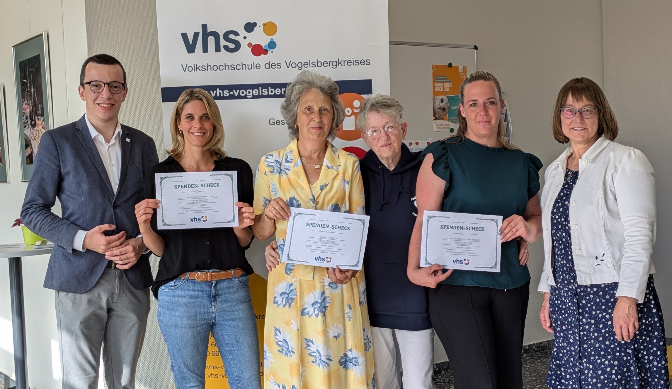 Sechs Menschen lächeln in die Kamera. Drei Vertreterinnen von Vereinen halten symbolische Checks über 100 Euro hoch, die der Erste Kreisbeigeordnete Patrick Krug gemeinsam mit vhs-Leiterin Monika Schenker übergeben hat.