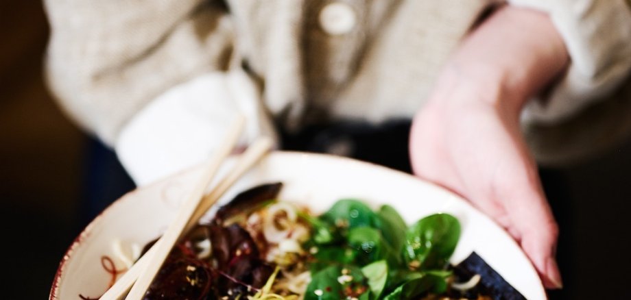 Eine Person hält einen Teller mit einer Ramensuppe in die Kamera. Zu sehen sind Sprossen, Eier, Nudeln und weitere Zutaten