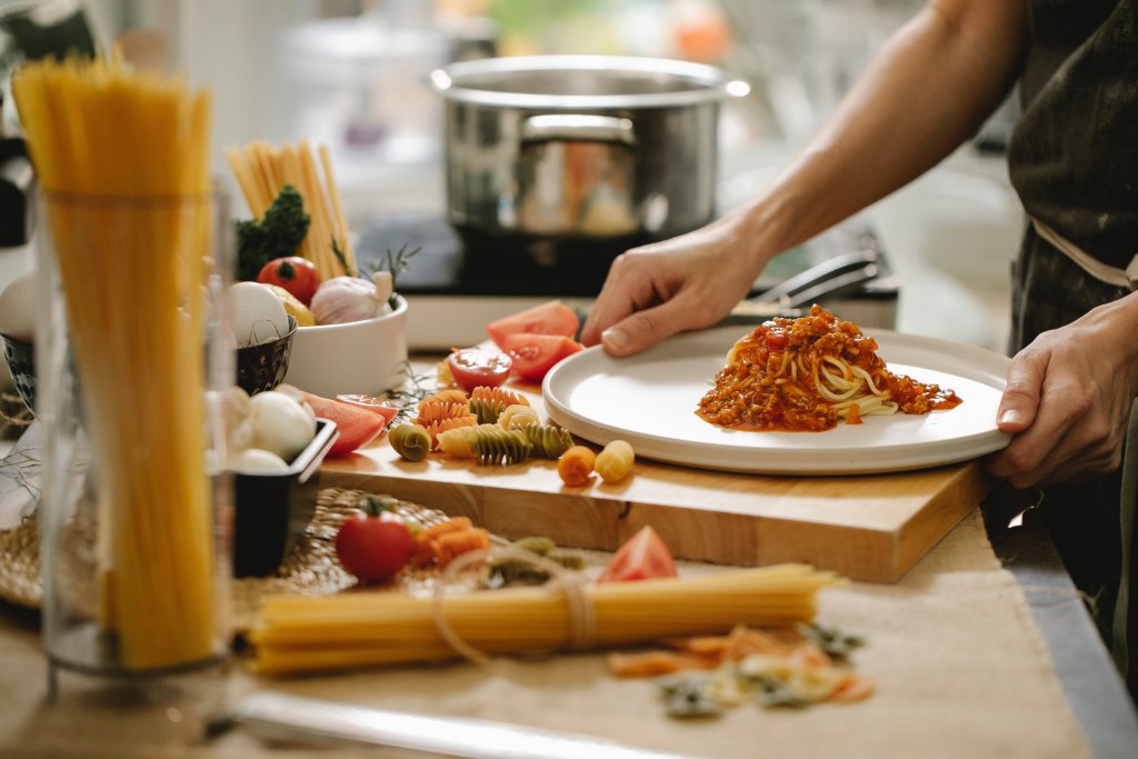Ein paar Hände sind darauf zu sehen, wie sie Spaghetti mit Tomatensauce zubereiten. Es liegen Nudeln auf einem Schneidebrett verstreut, im Hintergrund ist ein Top. 