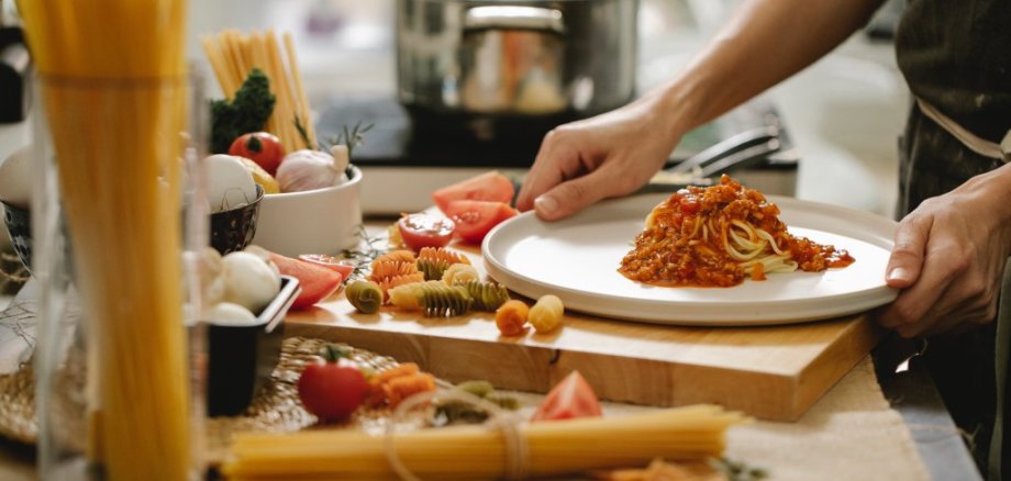 Ein paar Hände sind darauf zu sehen, wie sie Spaghetti mit Tomatensauce zubereiten. Es liegen Nudeln auf einem Schneidebrett verstreut, im Hintergrund ist ein Top.