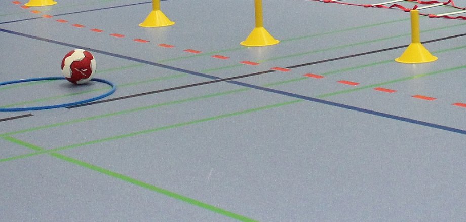 Nahaufnahme eines Sporthallenbodens. Darauf sind einige Hütchen, Markierungen und ein Handball zu sehen.