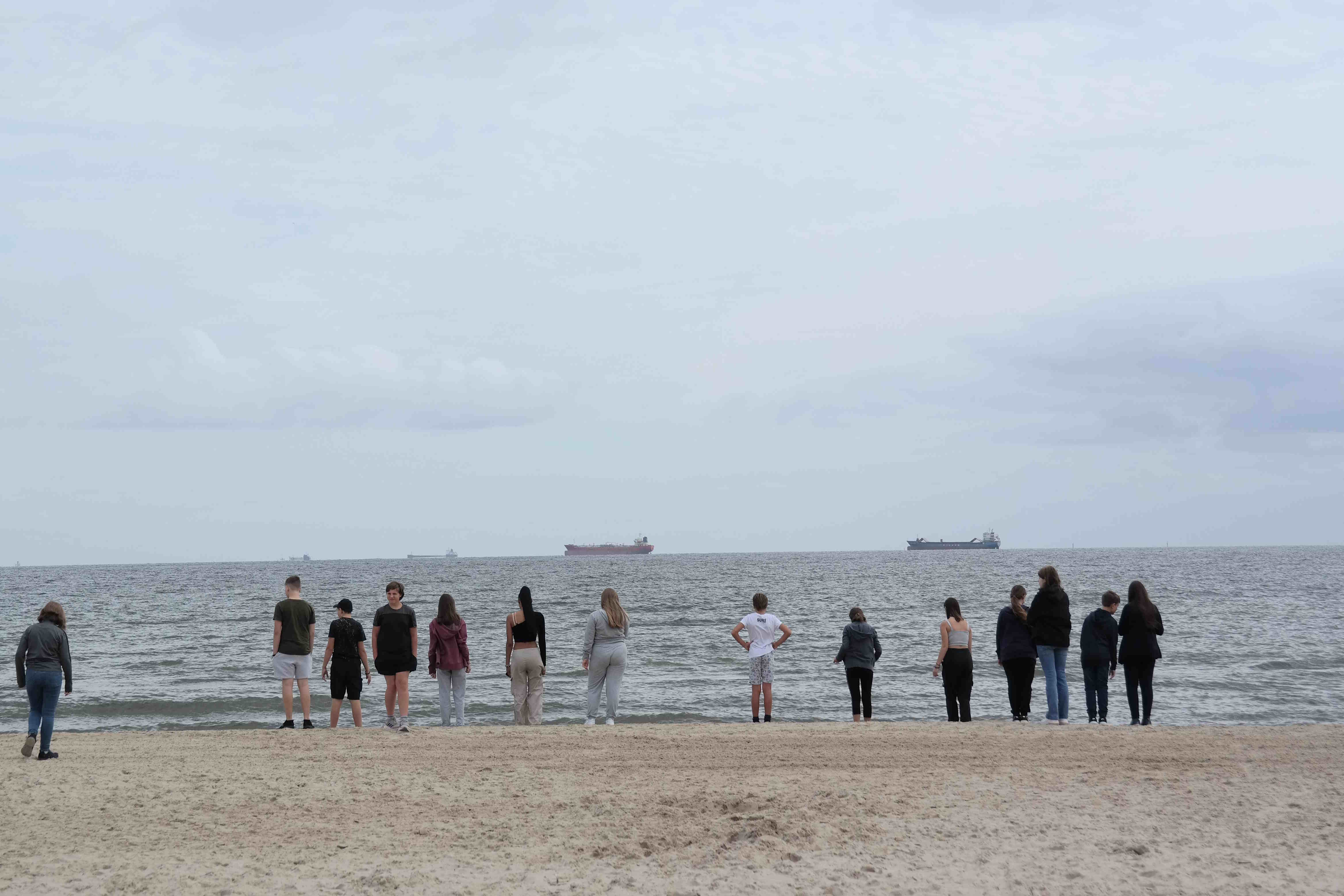 Eine Gruppe Jugendlicher steht am Strand von Utersum. Sie schauen in Richtung Meer. Im Hintergrund sind große Frachtschiffe zu sehen.