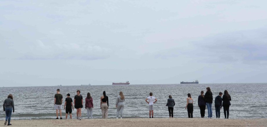 Eine Gruppe Jugendlicher steht am Strand von Utersum. Sie schauen in Richtung Meer. Im Hintergrund sind große Frachtschiffe zu sehen.