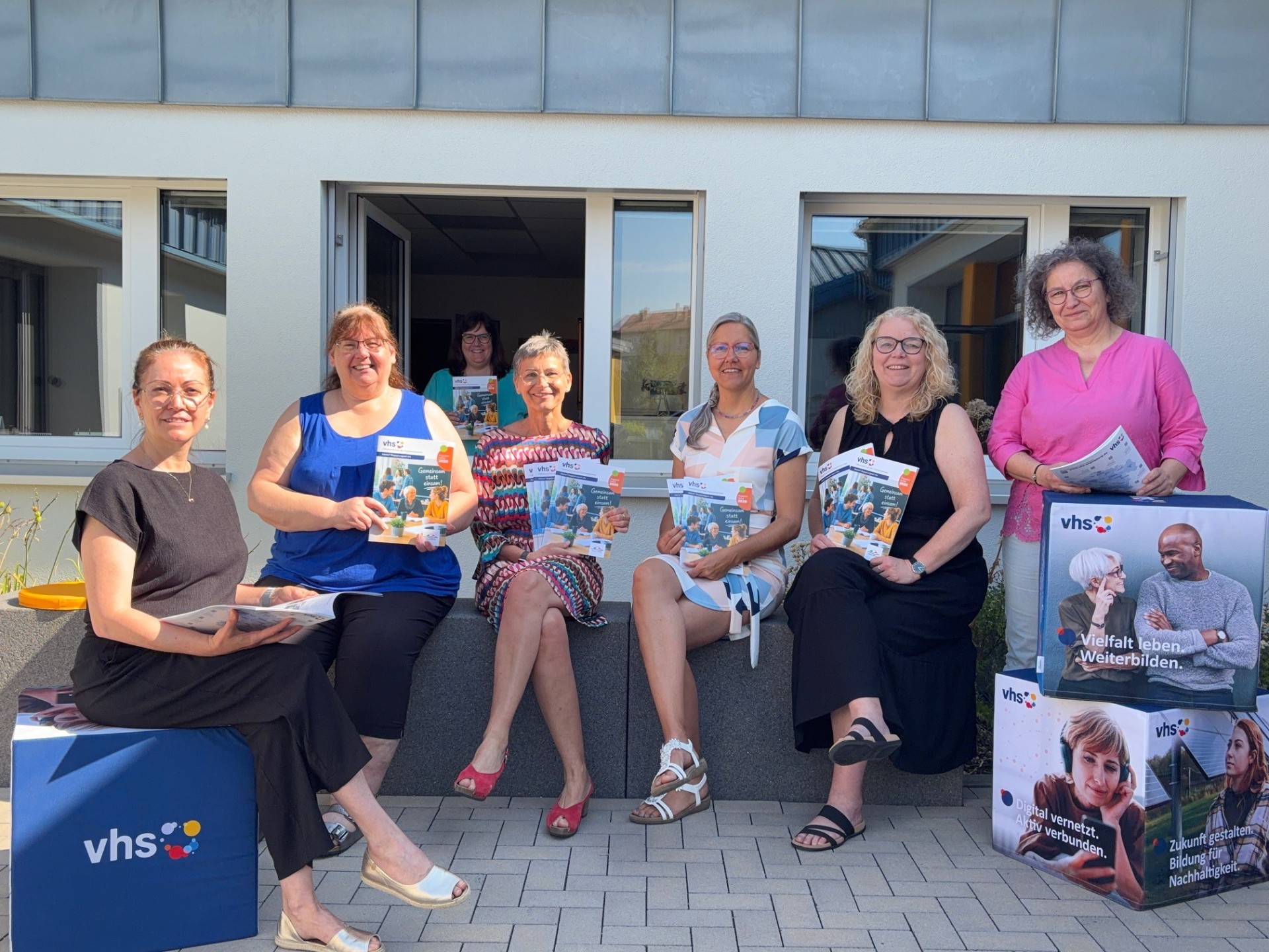Präsentiert das neue Programmheft: das Team der Volkshochschule mit vhs-Leiterin Kirsten Wegwerth (Mitte). Knapp zehn Menschen sitzen und stehen lässig neben einigen Aufstellern und halten das neue Programmheft in die Kamera.