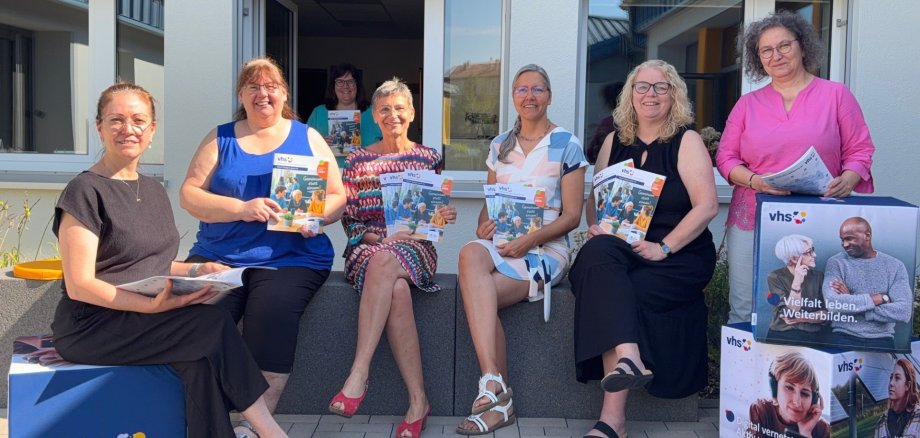 Präsentiert das neue Programmheft: das Team der Volkshochschule mit vhs-Leiterin Kirsten Wegwerth (Mitte). Knapp zehn Menschen sitzen und stehen lässig neben einigen Aufstellern und halten das neue Programmheft in die Kamera.
