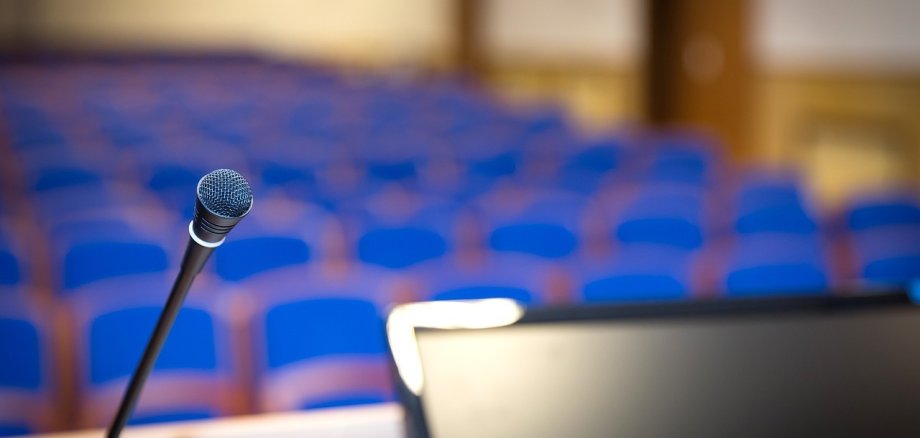 Blick über ein Rednerpult auf viele blaue Stuhlreihen. Die Ecke eines Bildschirms sowie ein Mikrofon sind zu sehen.