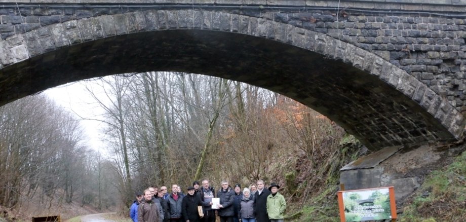 „Helft uns unsere Historische Brücke zu erhalten“ steht auf dem Schild – genau das tat Landrat Görig mit dem Überreichen einer OVAG-Spende an den Vereinsvorsitzenden Gerd Köhler. Grebenhains Bürgermeister Sebastian Stang, Mitglieder des Vereins „Historische Brücke Hartmannshain“ und des Ortsbeirats setzen sich dafür ein, dass die Brücke noch in diesem Jahr saniert und mit einem „Brückenfest“ wieder eröffnet werden kann. 