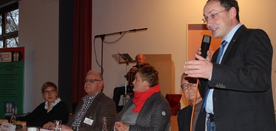Grebenhains Bürgermeister Sebastian Stang (stehend) konnte viele Besucherinnen und Besucher beim „Samstag für gesundes Altern“ begrüßen. Er steht am Ende des Tisches mit vier MEnschen, die alle an der Podiumsdiskussion teilnehmen. 