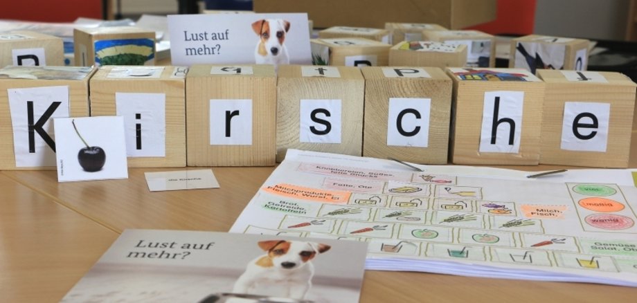 Symbolbild mit Materialien zum LEsenlernen. Darunter Holzklätze mit Buchstaben und Arbeitsblätter. Die Lerngruppe „Lesen und Schreiben von Anfang an“ trifft sich an zwei Vormittagen in der Woche: Außer Buchstaben und Wörtern wird heute gleichzeitig auch etwas zu gesunder Ernährung gelernt. 