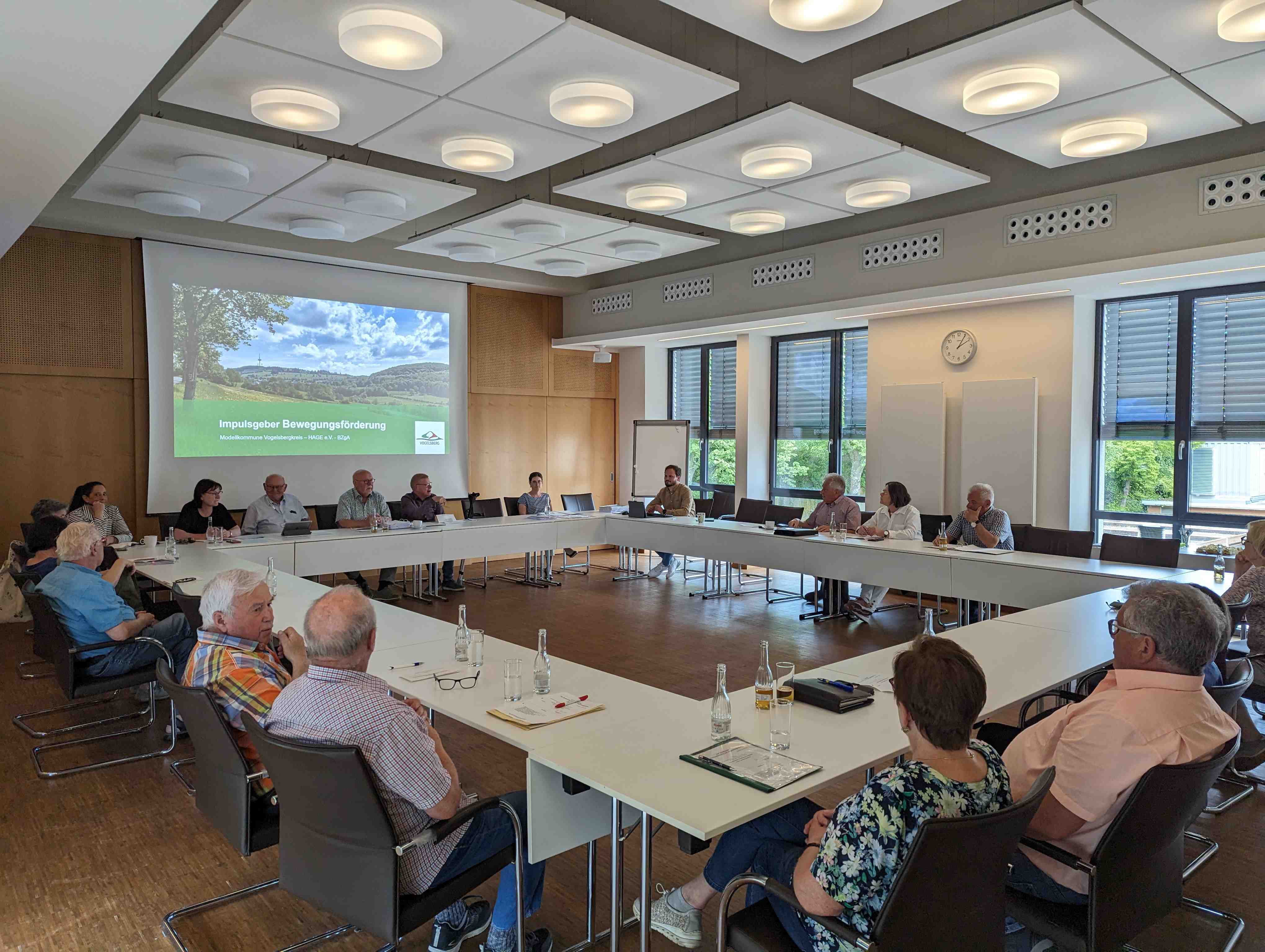 Eine Gruppe von Menschen sitzt in einem Sitzungssaal an Tischen verteilt. 
