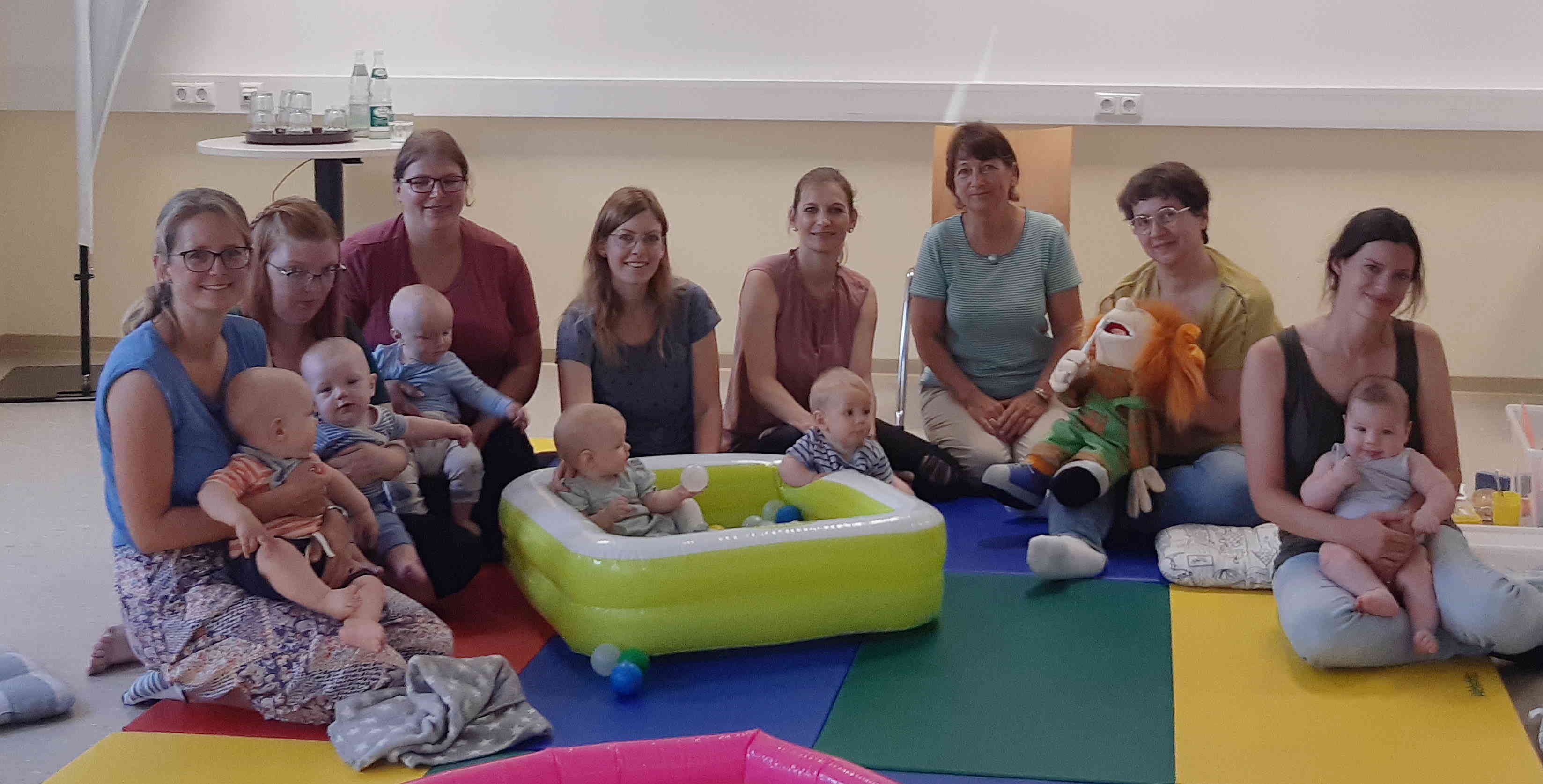 Eine Gruppe Mütter sitzt mit ihren Babys in einem Halbkreis. die Babys spielen auf einer bunten Matte und in einem Planschbecken.  