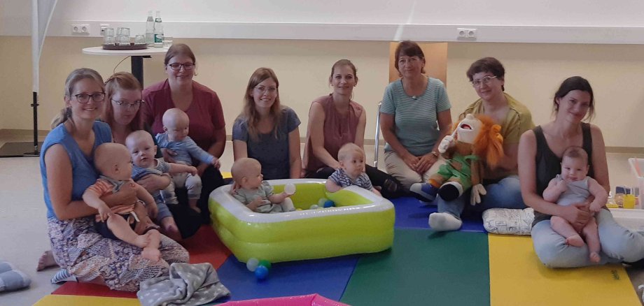 Eine Gruppe Mütter sitzt mit ihren Babys in einem Halbkreis. die Babys spielen auf einer bunten Matte und in einem Planschbecken.  