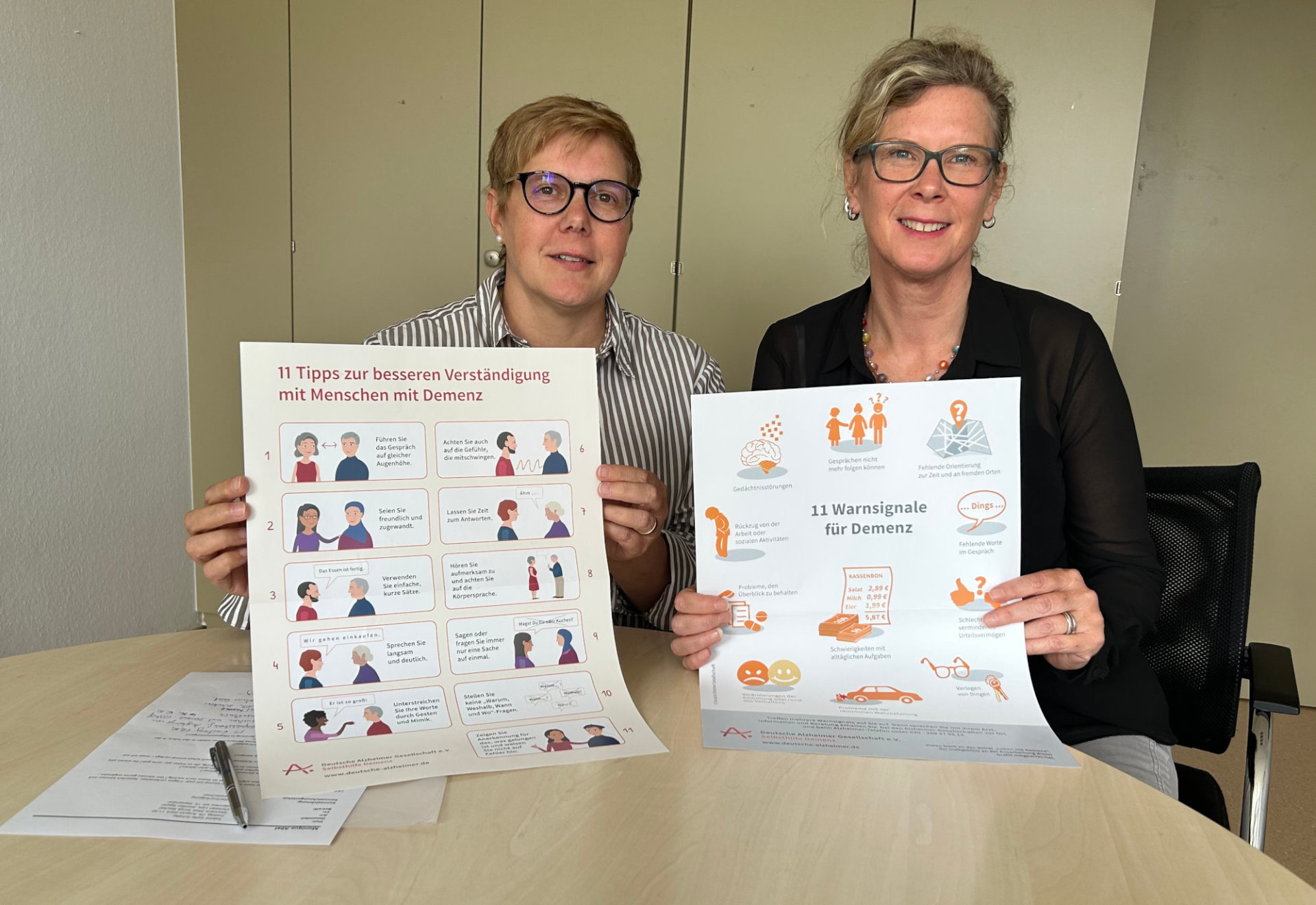 Zwei Frauen halten Plakate zum Thema Demenz in die Kamera und sitzen an einem Tisch.