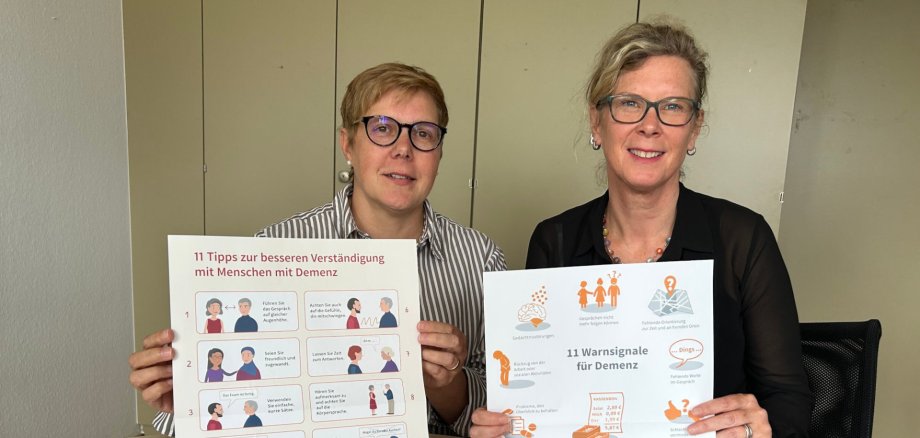 Zwei Frauen halten Plakate zum Thema Demenz in die Kamera und sitzen an einem Tisch.