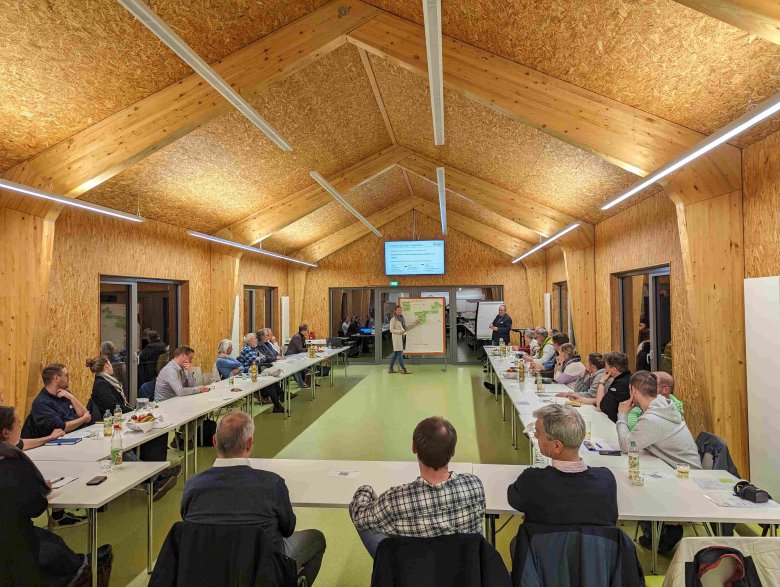 Abschließend werden die Ideen und Impulse aus den Kleingruppen im Plenum priorisiert. Etwa 30 Menschen sitzen in einem großen Raum und schauen zu einer Tafel