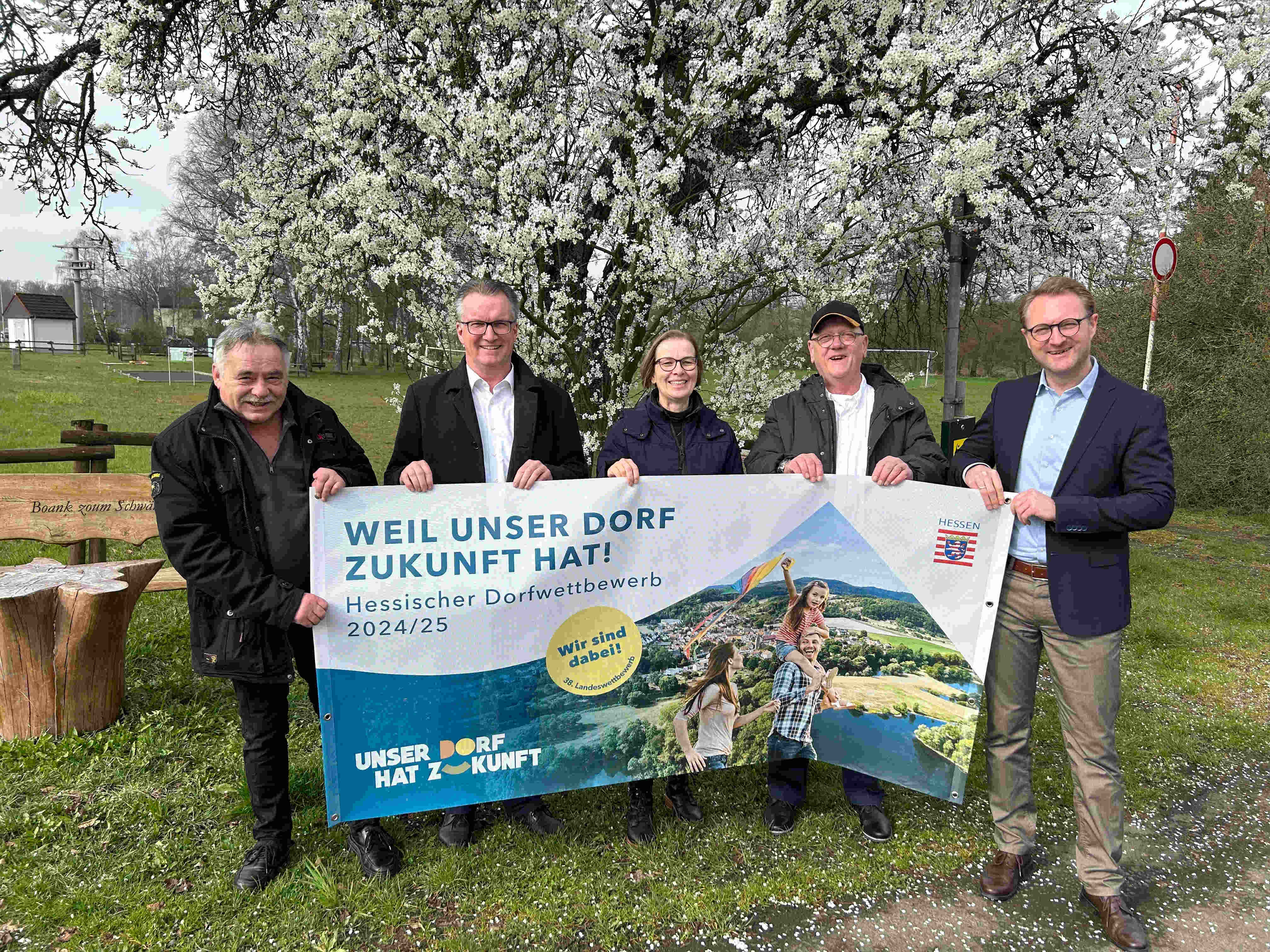 Vier Männer und eine Frau halten ein Banner hoch. Darauf wird für den Wettbewerb unser Dorf hat Zukunft geworben