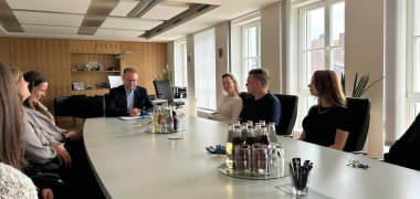 Mehrere Menschen sitzen an einem langen Konferenztisch und sind in eine entspannte Unterhaltung verwickelt