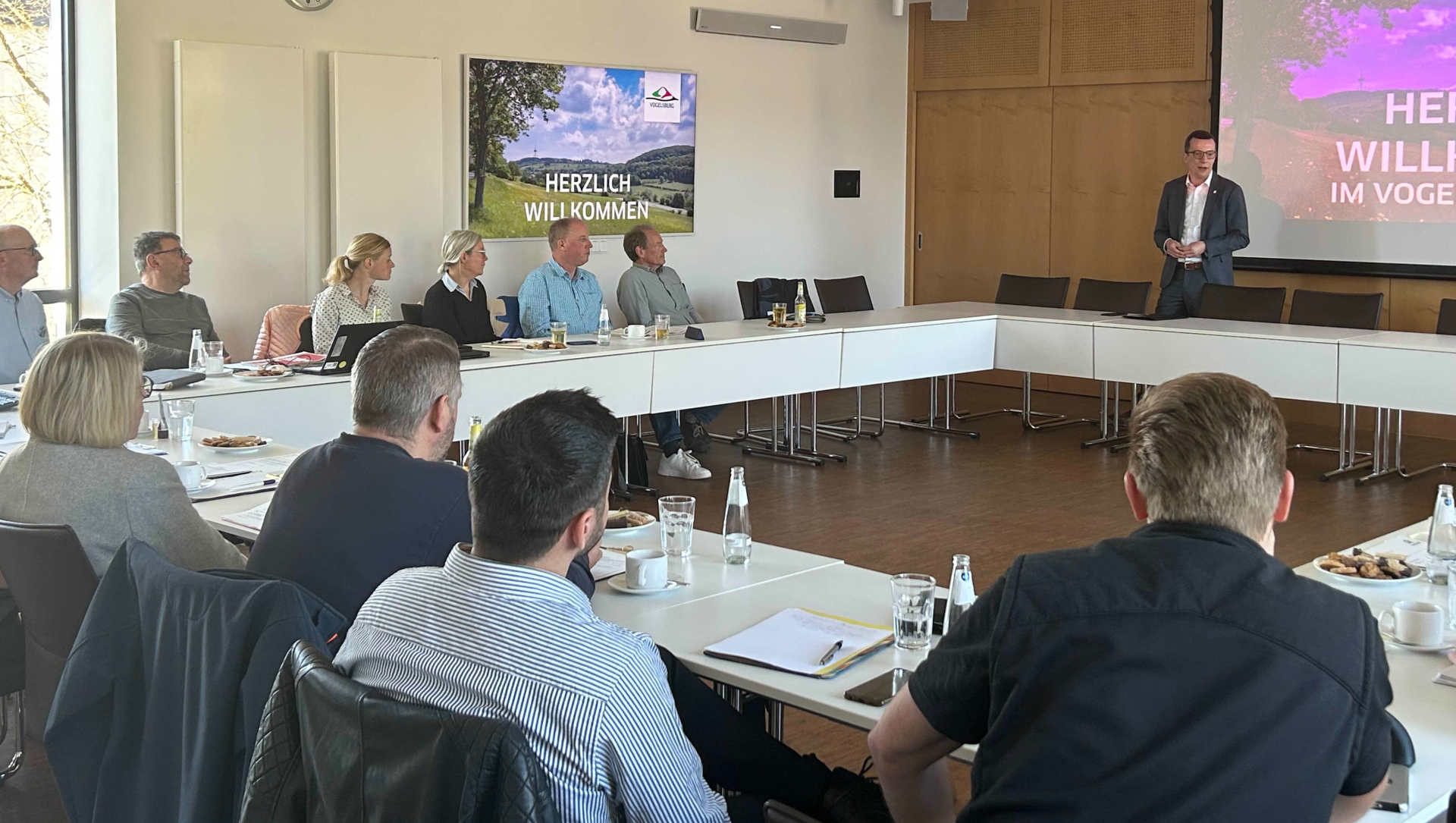 Eine Gruppe von Menschen sitzt in einem Sitzungssaal und hört einem Grußwort zu.