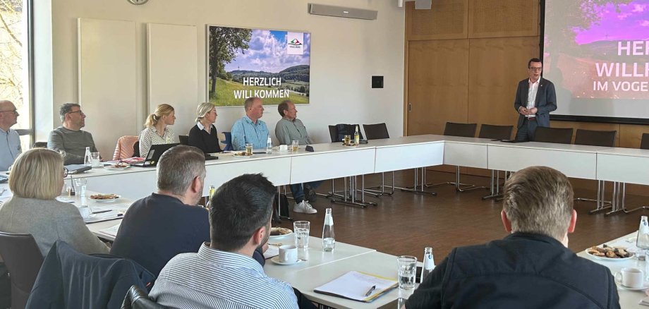 Eine Gruppe von Menschen sitzt in einem Sitzungssaal und hört einem Grußwort zu.