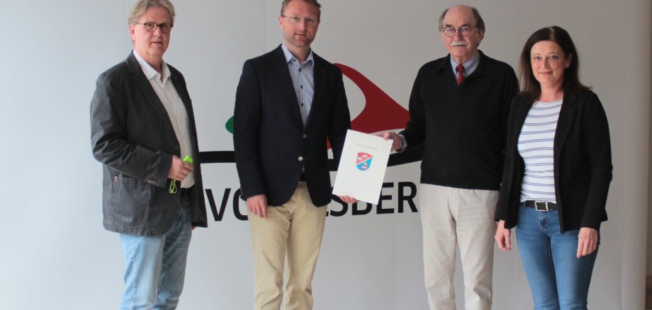 drei Männer und eine Frau stehen vor einer weißen Fotowand. Dr. Jens Mischak (Zweiter von links), Erster Kreisbeigeordneter und Jugenddezernent, übergibt den Zuwendungsbescheid an Rainer-Hans Vollmöller, Bürgermeister der Stadt Lauterbach, und Michaela Caspar-Merle, Sachgebietsleiterin für die Kindertagesstätten der Stadt Lauterbach. Helmut Benner (links), stellvertretender Leiter des Jugendamtes, ist federführend an der Entwicklung des Konzepts beteiligt.