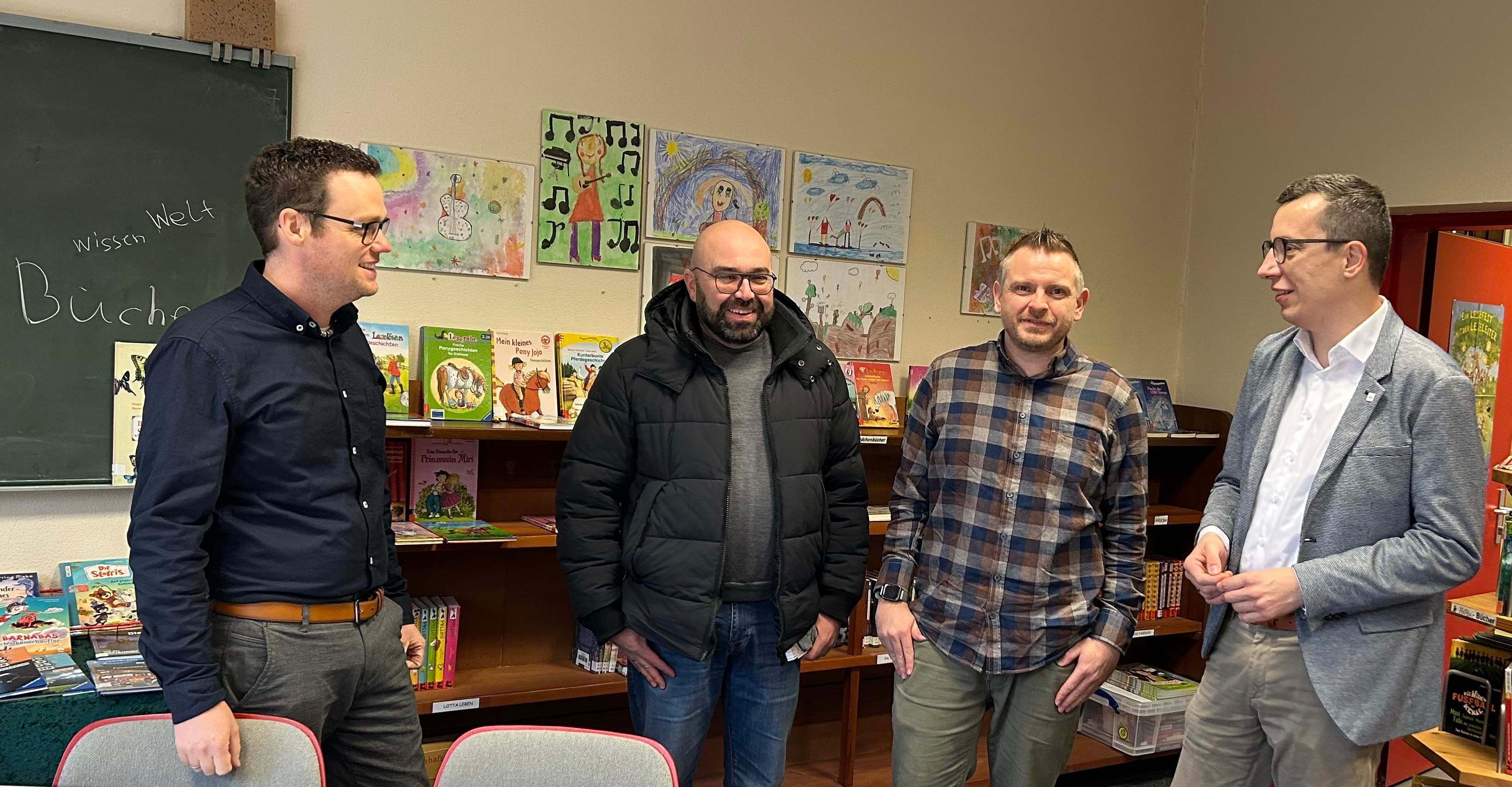 Erster Kreisbeigeordneter Patrick Krug im Austausch mit Schulleiter Nils Strowitzki, mit Stefan Schmidt, dem Leiter des Amtes für Hochbau, Energie und Gebäudewirtschaft, und Matthias Röse (von rechts), der das Amt für Schulische Bildung und Betreuung leitet. Sie stehen in einem Grundschulklassenraum mit Bildern der Schüler und einer Tafel im Hintergrund.