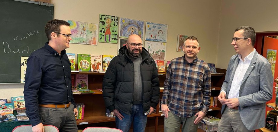 Erster Kreisbeigeordneter Patrick Krug im Austausch mit Schulleiter Nils Strowitzki, mit Stefan Schmidt, dem Leiter des Amtes für Hochbau, Energie und Gebäudewirtschaft, und Matthias Röse (von rechts), der das Amt für Schulische Bildung und Betreuung leitet. Sie stehen in einem Grundschulklassenraum mit Bildern der Schüler und einer Tafel im Hintergrund.
