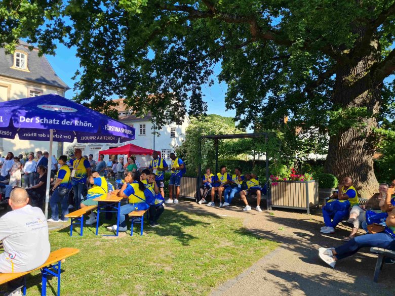 Besucher eines Fests sitzen unter Schirmen und einem Baum an Bierzeltgarnituren. 