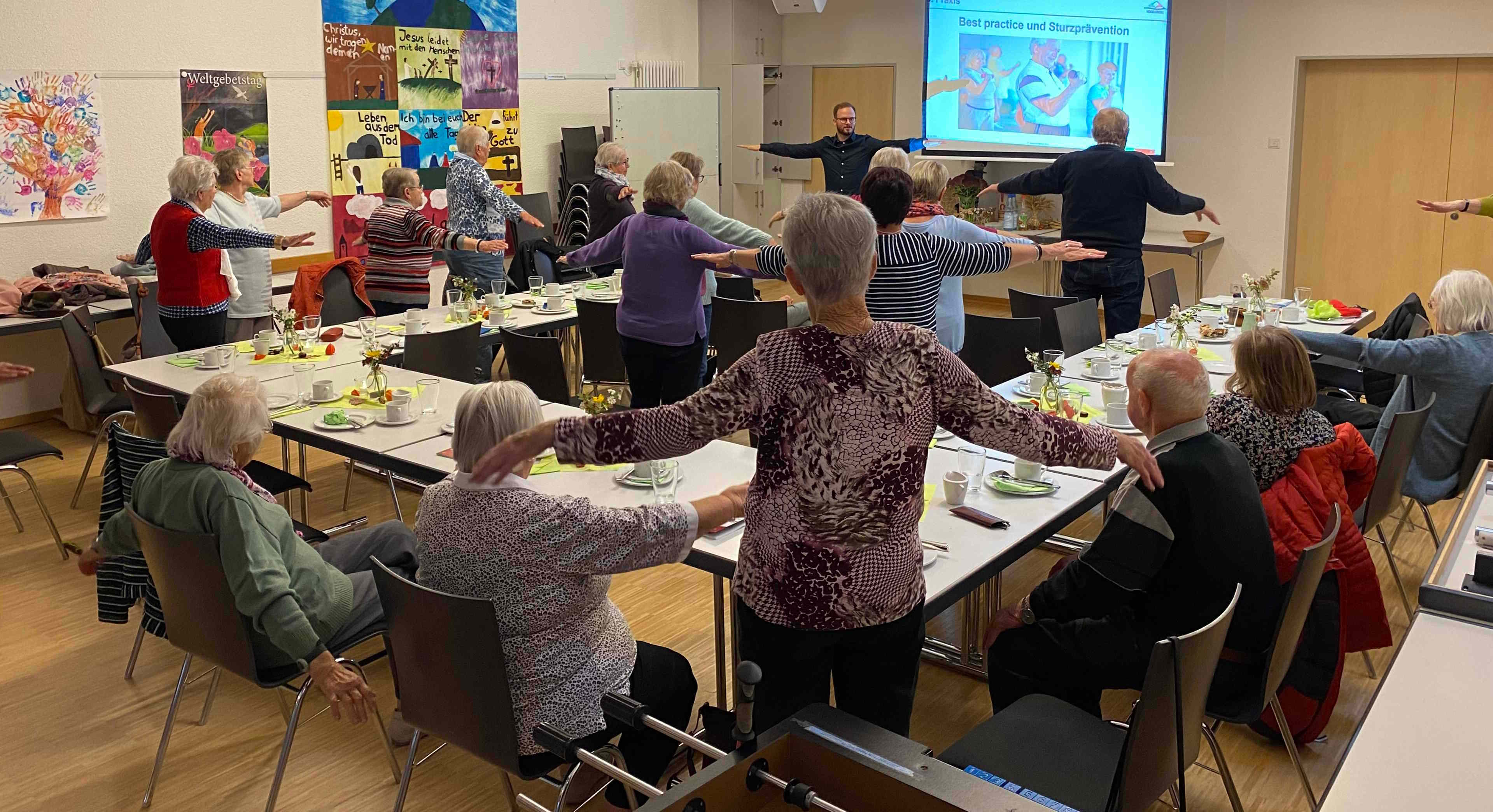 Eine Gruppe von Senioren macht gemeinsam mit Dominic Günther Koordinations- und Bewegungsübungen in einem Gemeindesaal