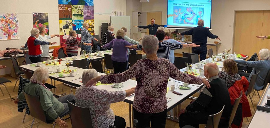 Eine Gruppe von Senioren macht gemeinsam mit Dominic Günther Koordinations- und Bewegungsübungen in einem Gemeindesaal