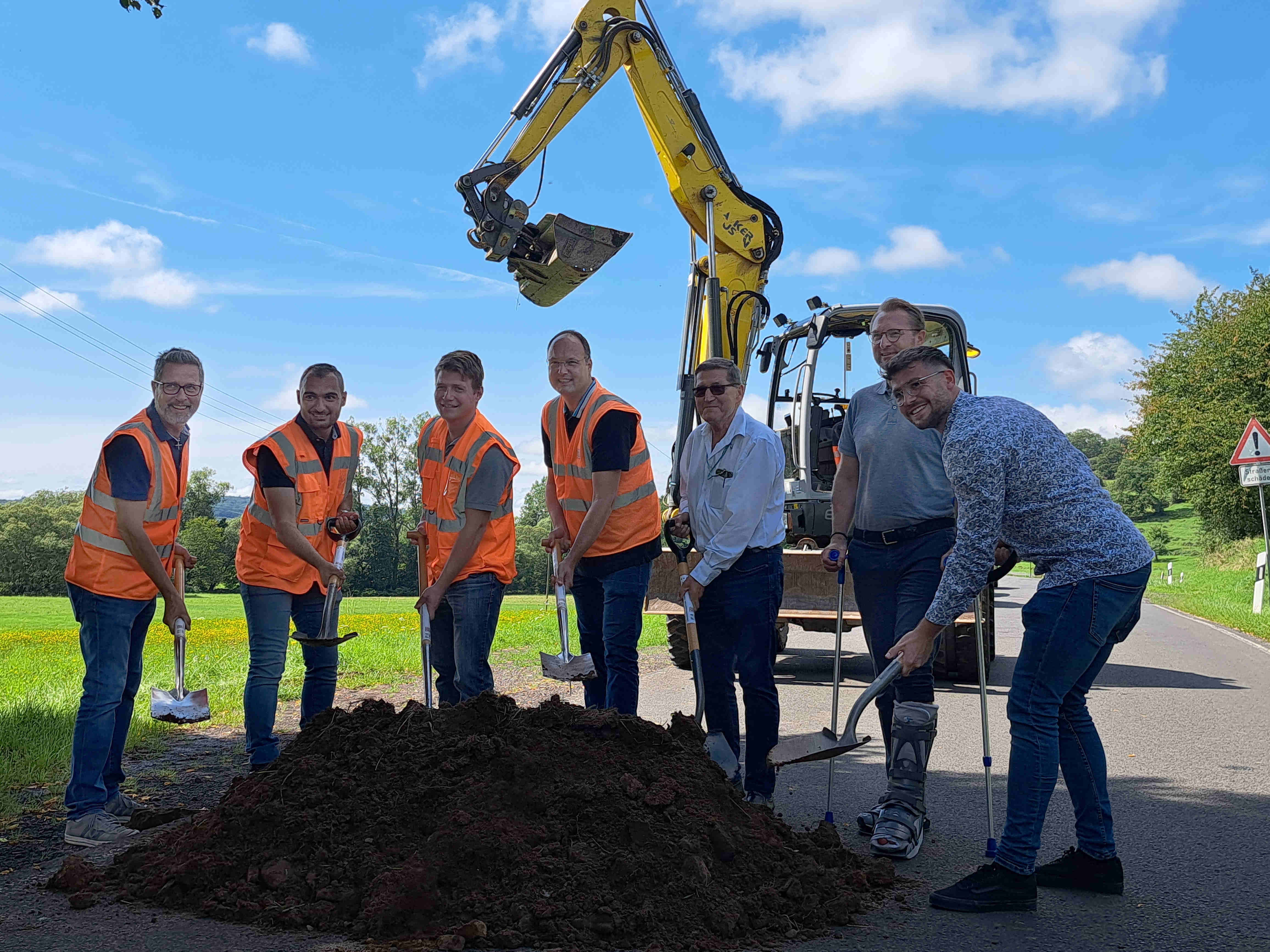 Sieben Männer stehen mit Warnwesten und Spaten an einem Erdhaufen für einen symbolischen Spatenstich. Im Hintergrund ist ein Bagger zu sehen.