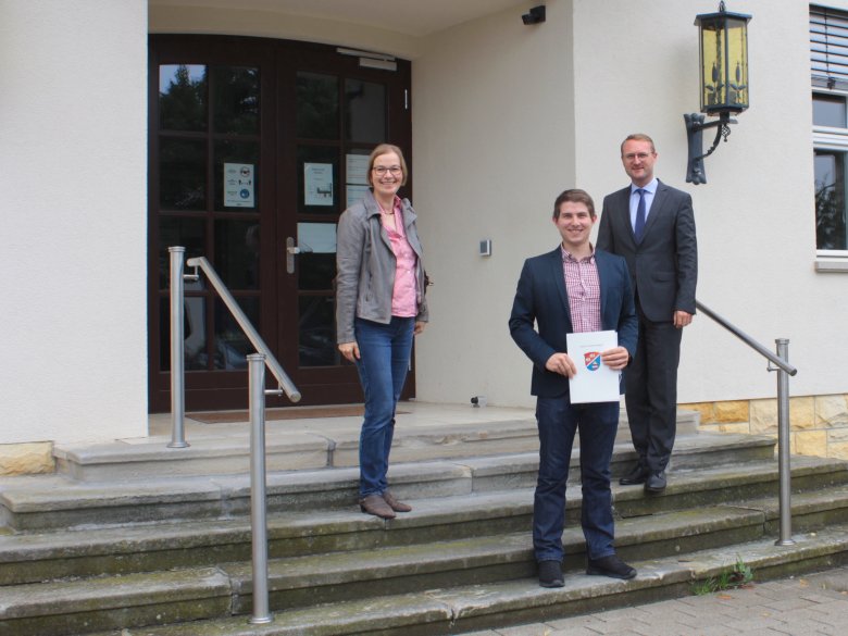 Florian Rausch zeigt den Förderbescheid, den Dr. Mischak und Angelika Boese gerade überreicht haben. Sie stehen vor dem Eingang zur Kreisverwaltung auf einer Treppe.