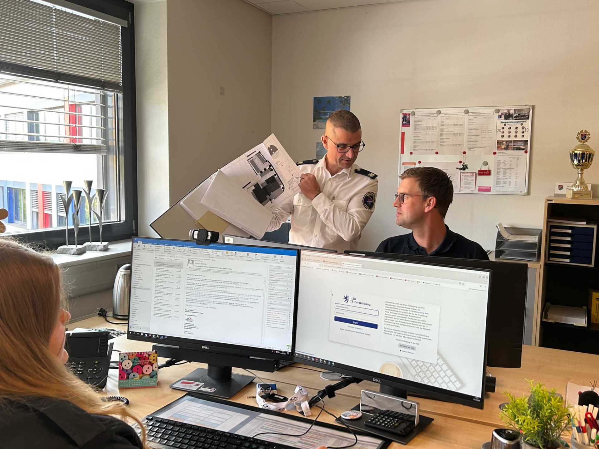 Drei Menschen stehen und sitzen in einem Büro und präsentieren einen Bauplan. 