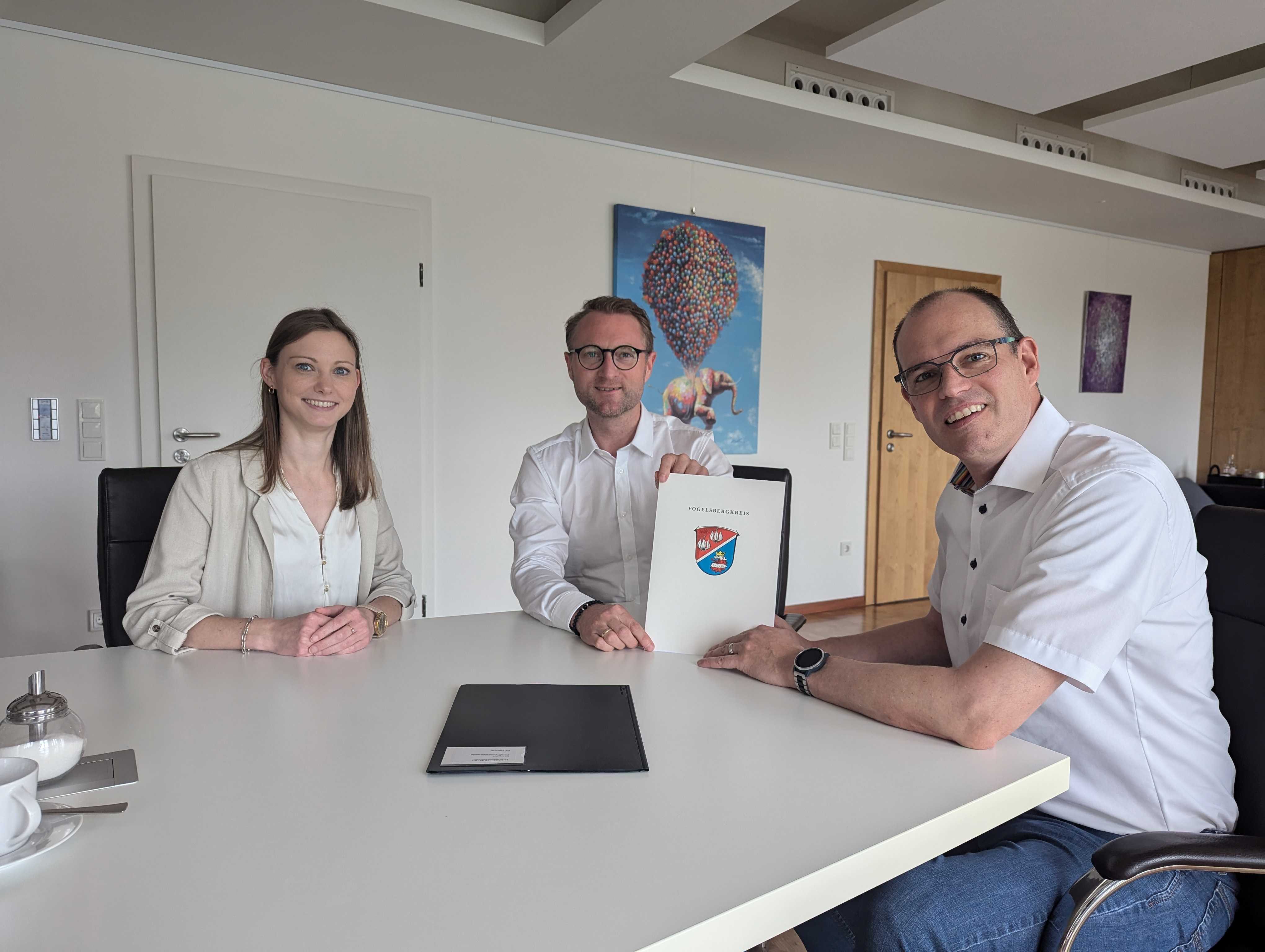 Landrat Dr. Jens Mischak (Mitte) übergibt gemeinsam mit Jana Brittner, vom Amt für Wirtschaft und den ländlichen Raum des Vogelsbergkreises, drei Dorfentwicklungs-Förderbescheide an Sascha Spielberger, Bürgermeister der Gemeinde Freiensteinau. Sie sitzen an einem Tisch und präsentieren die Bescheide