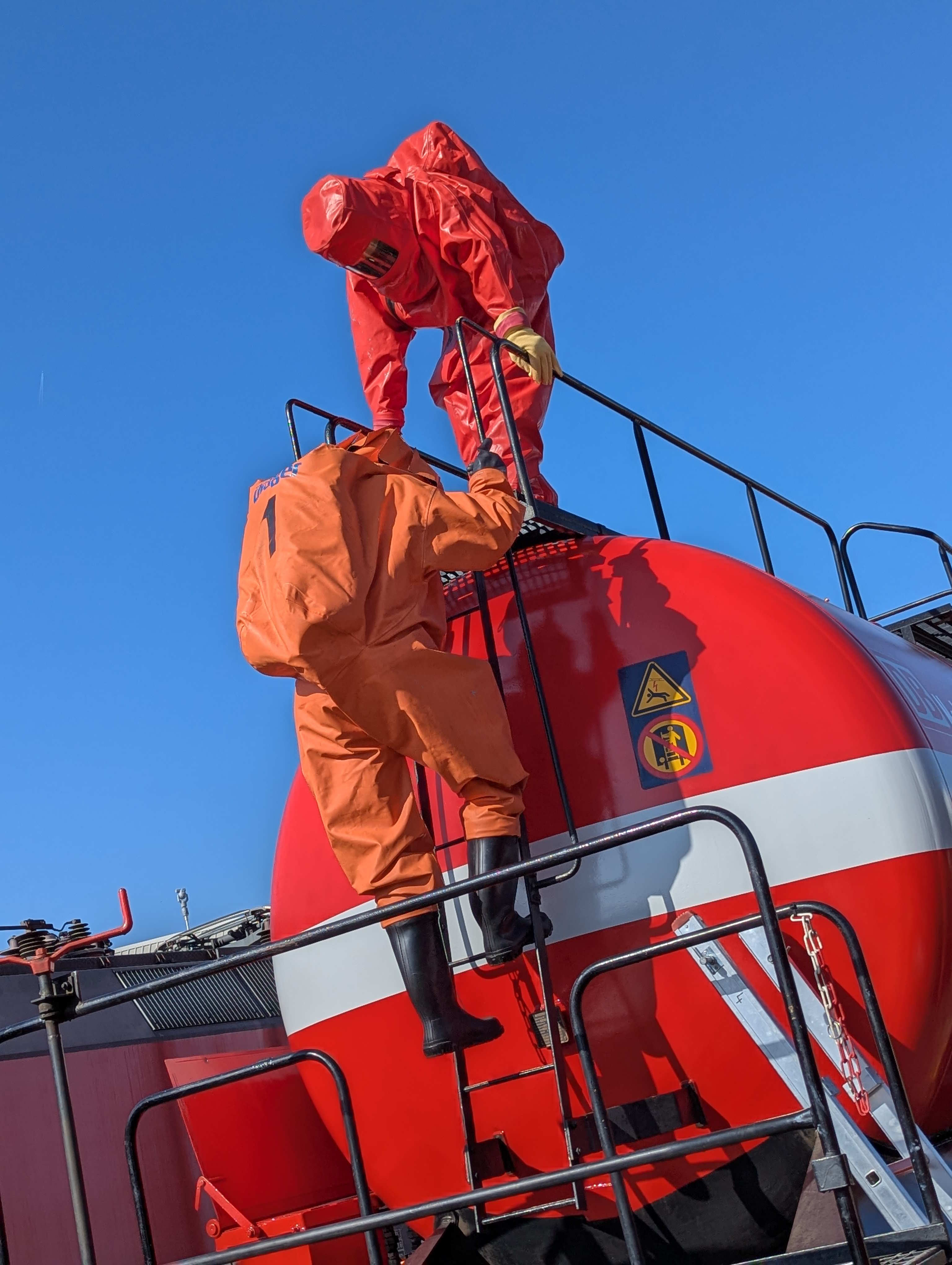 Ein Feuerwehrmann klettert auf einen roten Tankwaggon. Er trägt einen Gefahrstoffschutzanzug in orange. 