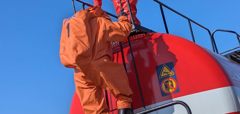 Ein Feuerwehrmann klettert auf einen roten Tankwaggon. Er trägt einen Gefahrstoffschutzanzug in orange. 