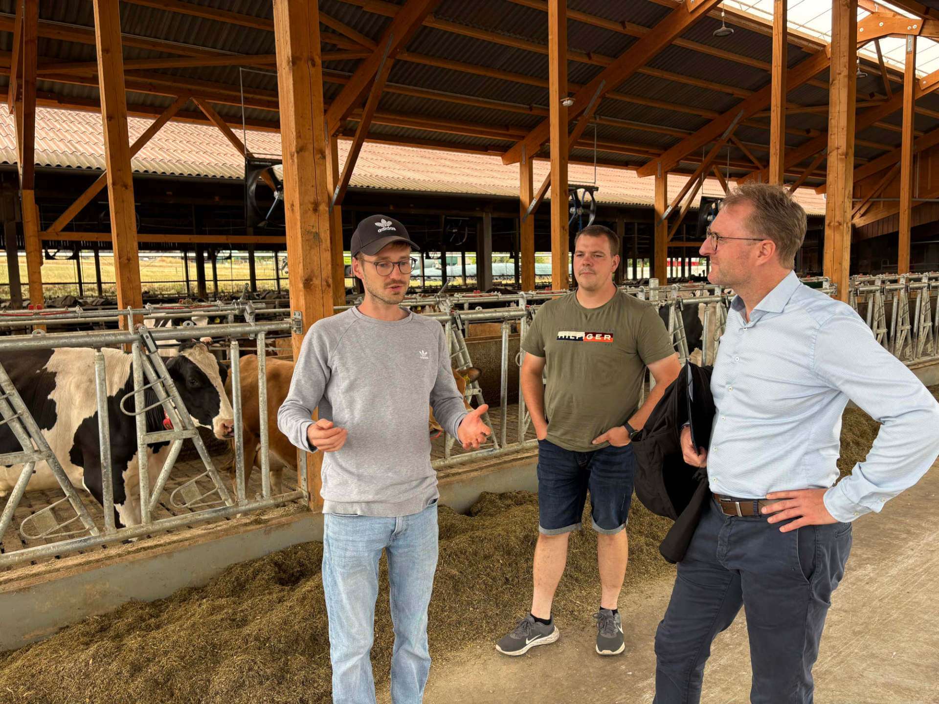 Herausfordernde Zeiten: Dennis und Cousin Aaron Semmler (links und Mitte) beschrieben Dr. Jens Mischak (rechts), inwiefern hohe Betriebskosten sowie die anhaltende Trockenheit die Arbeit auf dem Hof beeinflussen. Sie stehen in einem hellen Kuhstall und sind im Gespräch.