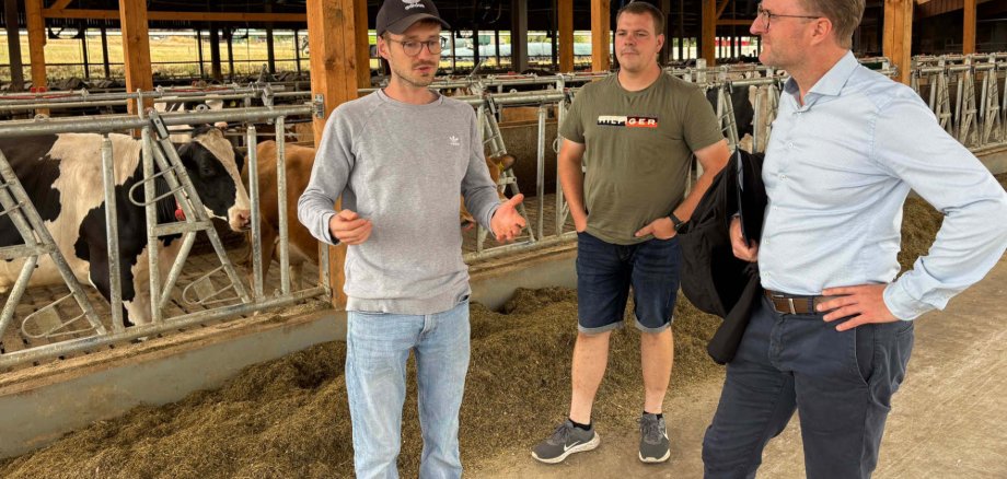 Herausfordernde Zeiten: Dennis und Cousin Aaron Semmler (links und Mitte) beschrieben Dr. Jens Mischak (rechts), inwiefern hohe Betriebskosten sowie die anhaltende Trockenheit die Arbeit auf dem Hof beeinflussen. Sie stehen in einem hellen Kuhstall und sind im Gespräch.