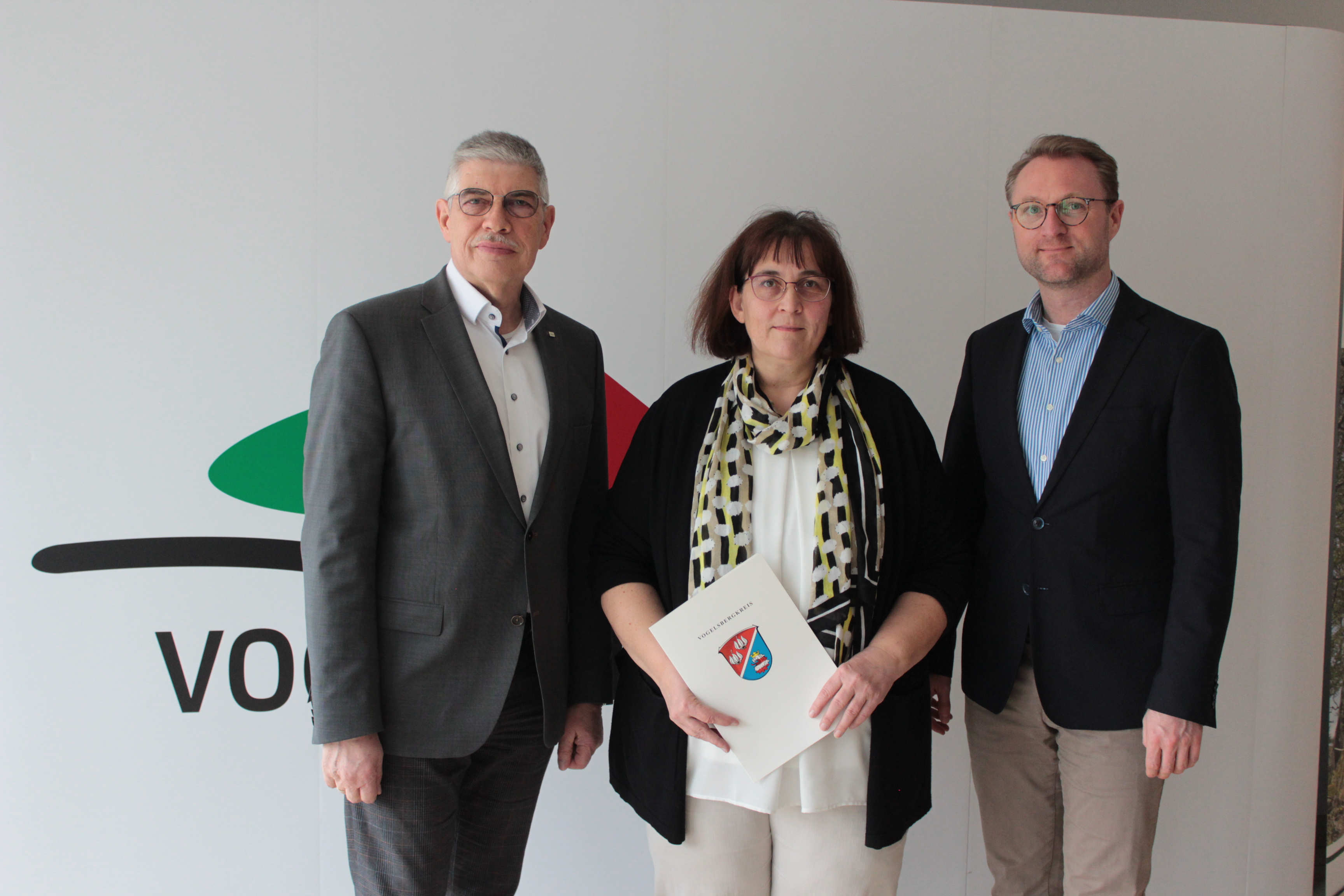 Landrat Manfred Görig und Erster Kreisbeigeordneter Dr. Mischak gratulieren Andrea Schönfeld zum 25-jährigen Dienstjubiläum. Sie stehen vor einer weißen Fotowand. 