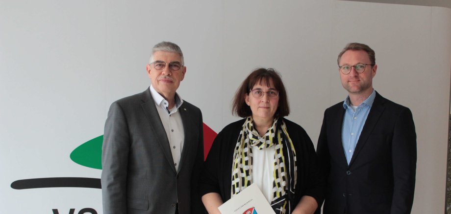 Landrat Manfred Görig und Erster Kreisbeigeordneter Dr. Mischak gratulieren Andrea Schönfeld zum 25-jährigen Dienstjubiläum. Sie stehen vor einer weißen Fotowand. 