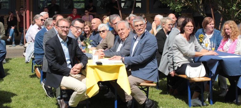Blick auf die Ehrengäste. Sie sitzen an Bierzeltgarnituren bei schönem Wetter im Innenhof einer Schule.