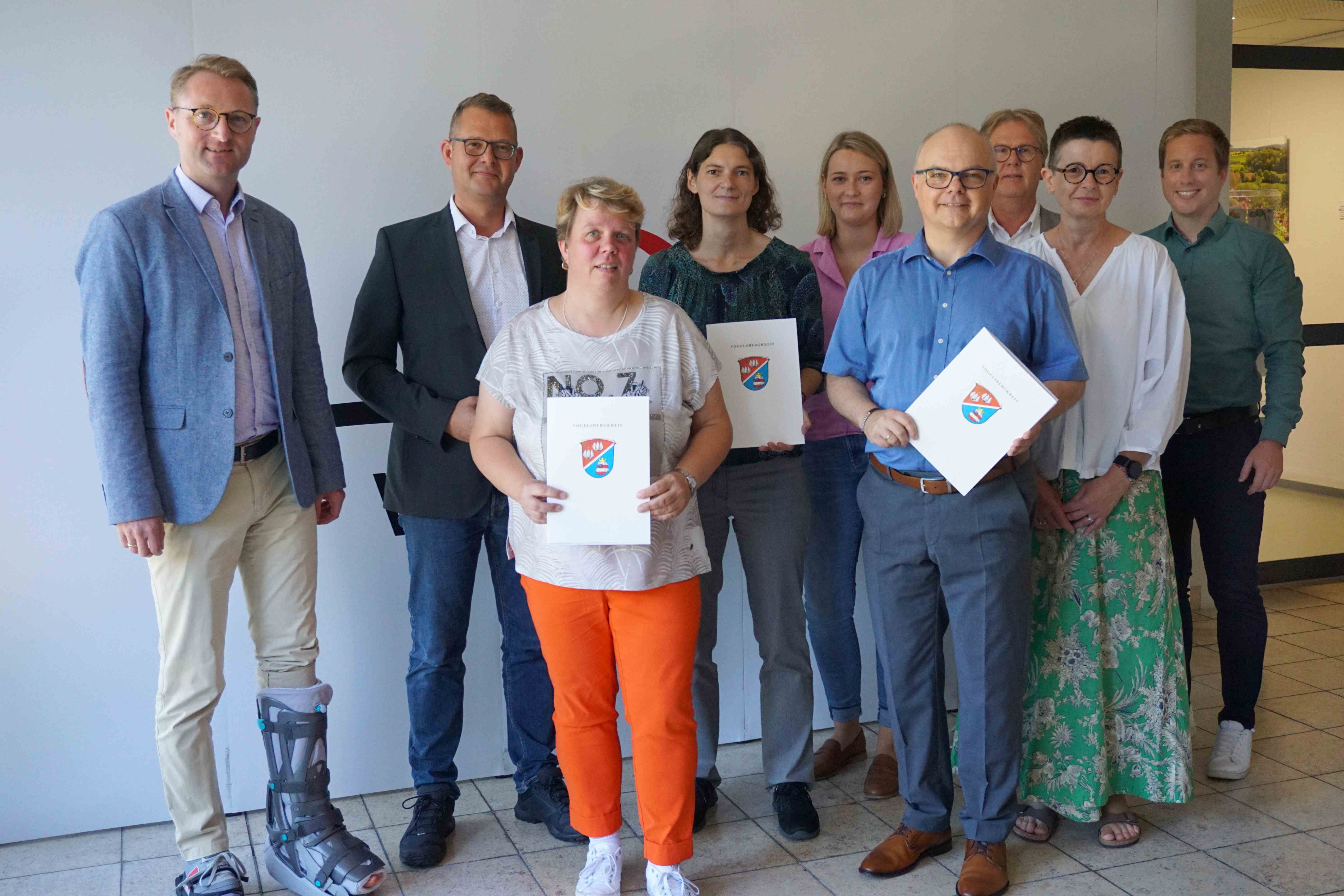 Mehr Planungssicherheit für vier Vogelsberger Kitas: Dr. Jens Mischak steht gemeinsam mit acht weiteren Menschen vor einer weißen Fotowand. Er hat Bescheide zur Schaffung inklusiver Kita-Plätze übergeben. 