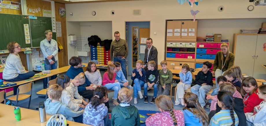 Eine Grundschulklasse sitzt in einem Stuhlkreis und spricht mit einem Lehrer. Im Hintergrund schauen sich vier Erwachsene die Unterrichtsstunde an. 