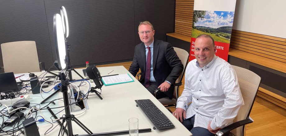 Erster Kreisbeigeordneter Dr. Jens Mischak (links) und der Referent für Elektromobilität Stefan Hirth (OVAG) während der Online-Abendveranstaltung. Sie sitzen vor Streaming-Equipment. Eine Lampe, ein Mikrofon und eine Kamera sind aufgebaut
