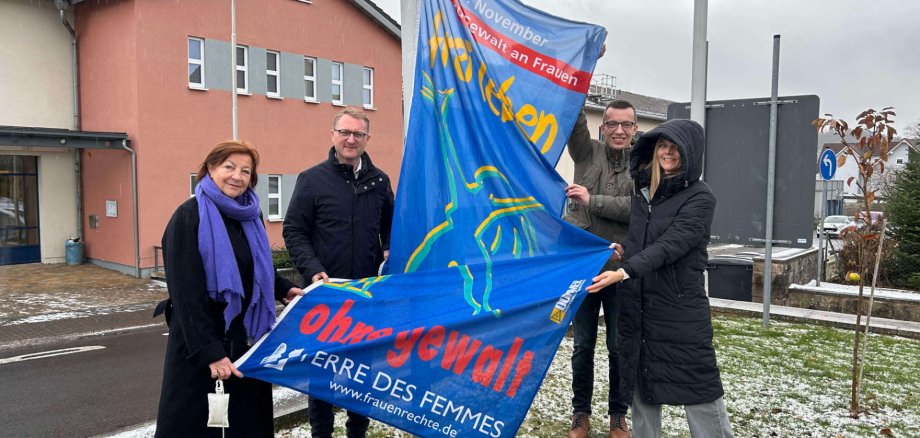Vier Personen hissen symbolisch eine Flagge, die zum Schutz von Frauen gegen Gewalt aufruft.
