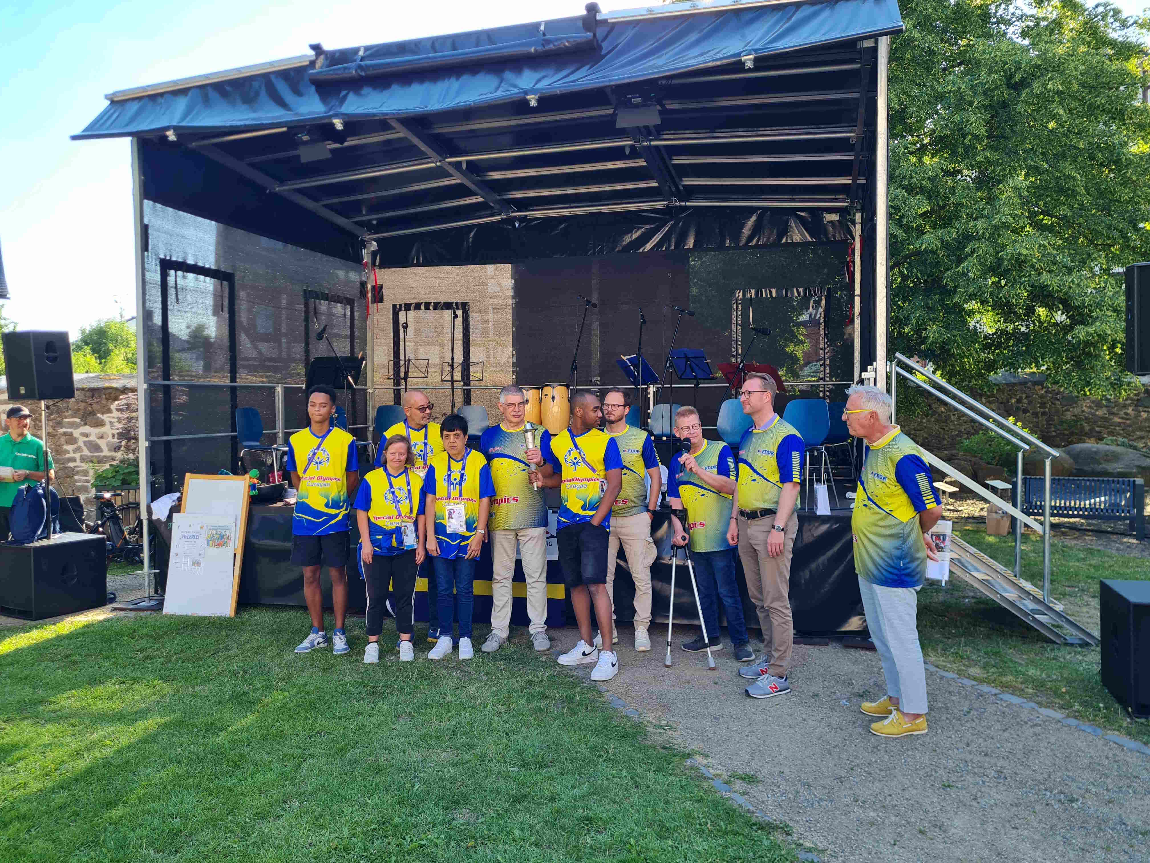 Vor einer Bühne stehen 10 Personen in Gelb-Blauen Trikots. Zwei halten eine brennende Fackel.