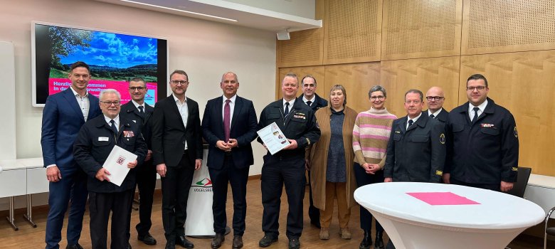 Zwölf Menschen haben sich in einem Sitzungssaal zum Gruppenbild aufgestellt. Sie lächeln in die Kamera und präsentieren die Auszeichnung für den Vogelsbergkreis als "Feuerwehr des Monats"