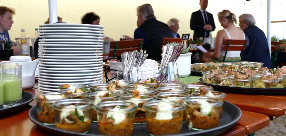 Auf einem Tisch ist Essen in kleinen Gläschen zu sehen. Im Hintergrund sitzen Menschen an einem Tisch und Sprechen